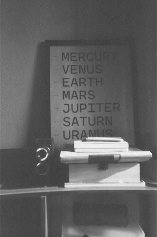 a camera, a wooden box, a couple of notebooks, and a poster listing the names of planets perched on a desk shelf